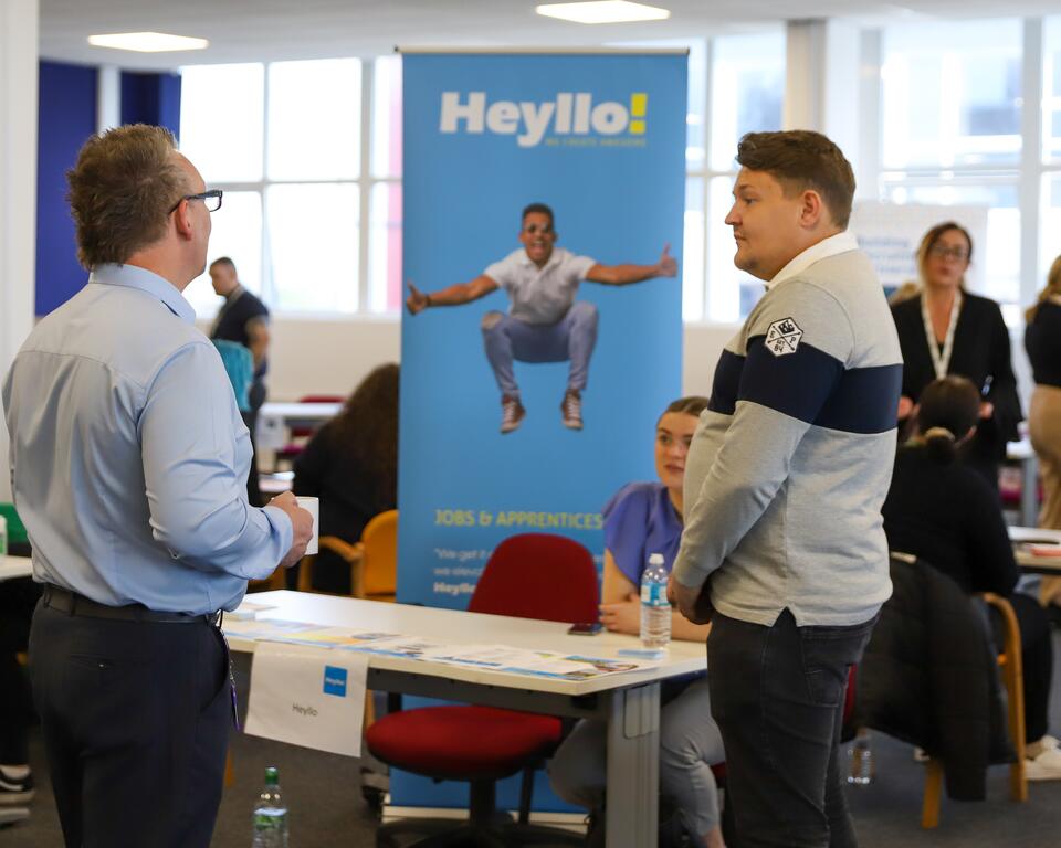 Two men chatting in front of a desk in a busy office.