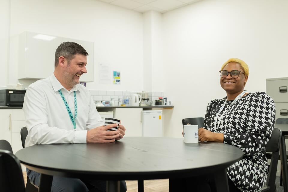 Man and woman chatting in an office kitchen while sitting at a round table, both having a mug of coffee.
