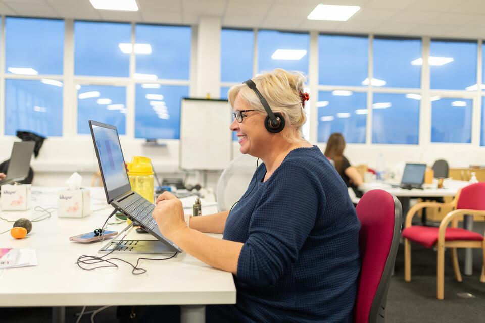 Smiley, blonde woman wearing a headset typing on her laptop at her desk in a busy office.