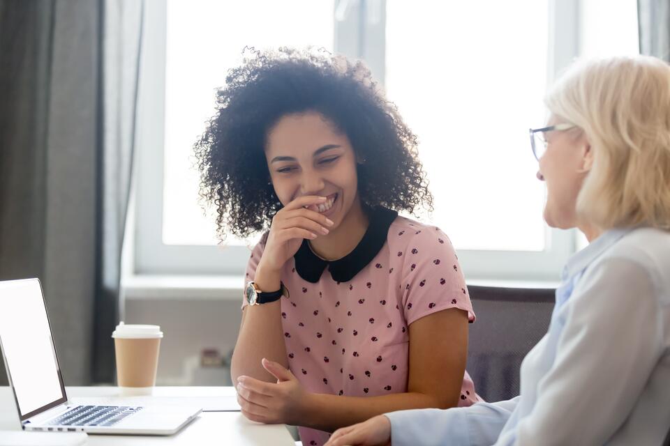 Young woman smiling at older woman with a coffee and laptop on the desk in front of them,