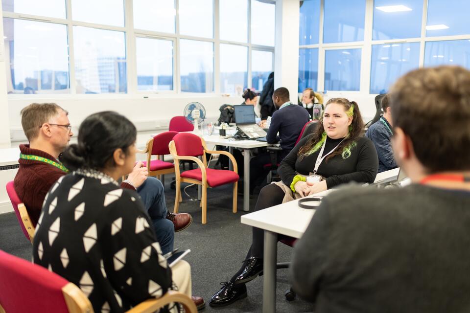 Group of four people sitting in a busy office with three listening intently to a woman who is speaking. 