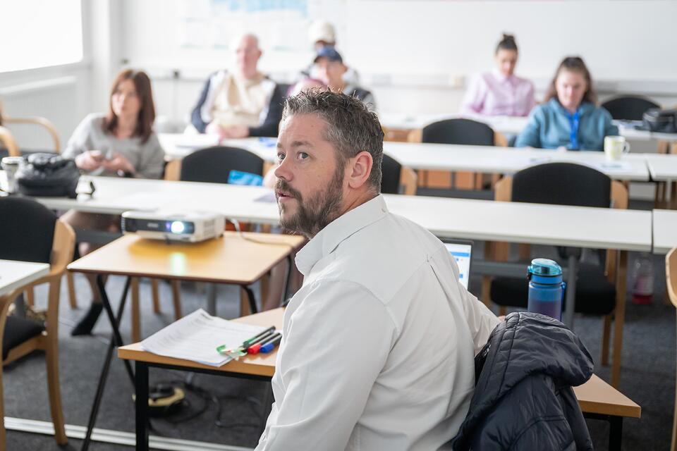 Man teaching at front of room with audience at desks behind him with everyone including teacher, looking at something at the front of the room. 