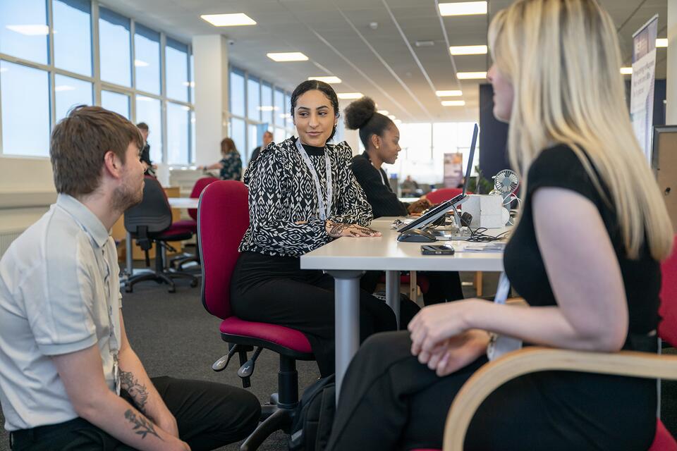 Three people sitting at a desk chatting. Two women sit in chairs while a man kneels beside them talking.