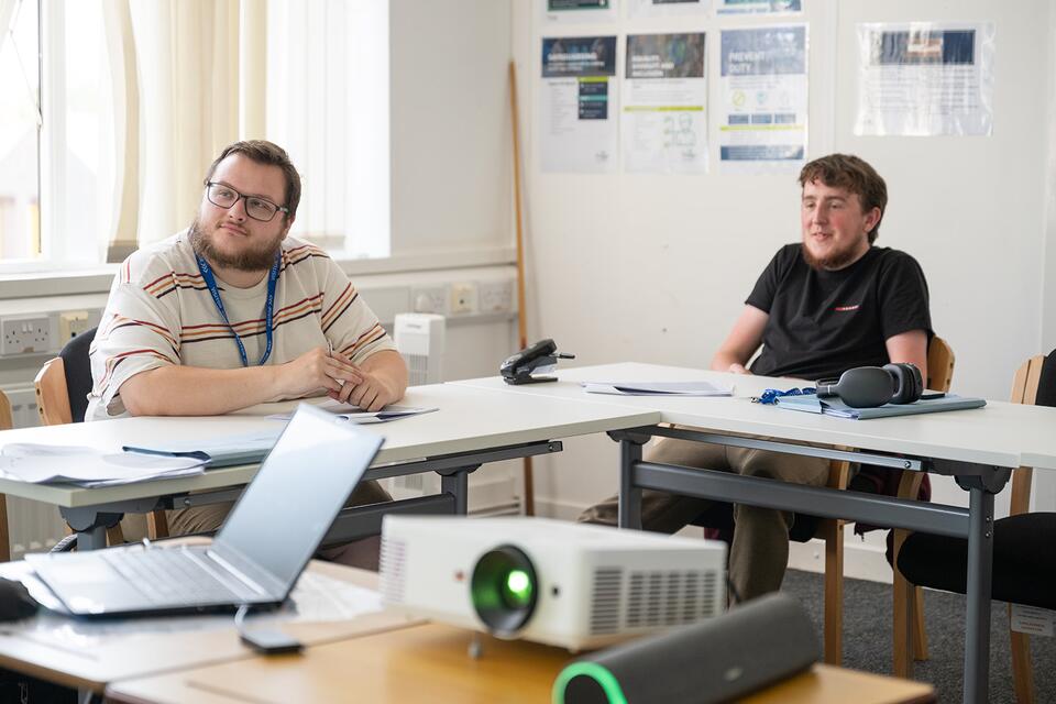 Two people sitting in a classroom style room at desks looking towards the front of the room (at tutor).