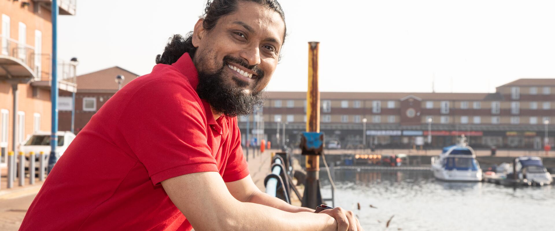 Man in red t-shirt posing by water with boats.