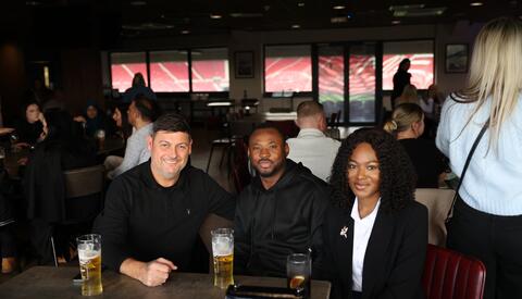 Two men and one woman sat at a table, with beer and coke, smiling for the camera. In the background, the event room/ bar is busy with colleagues smiling and laughing.