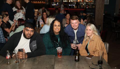 Two men, two women sit at a table in a bar posing for camera. on the table are drinks.