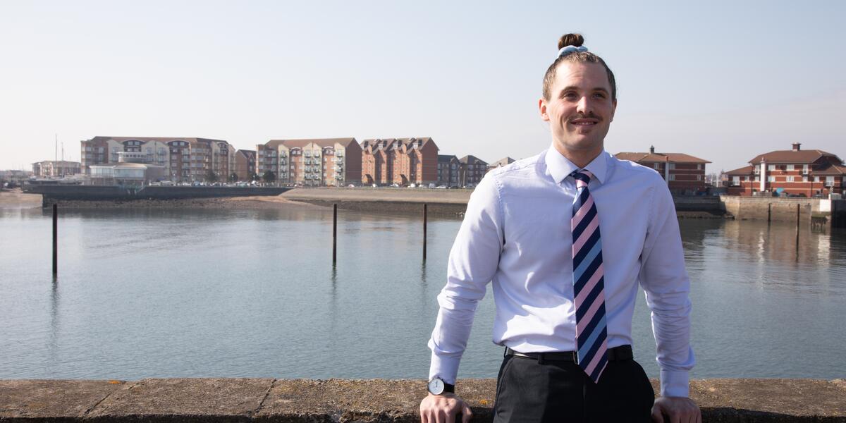 Man in shirt and tie smiling at the camera with water and buildings in background.