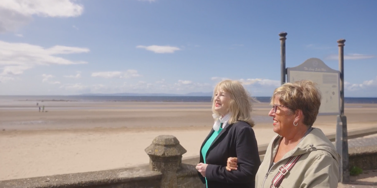 Two women walking and smiling with beach in the background.