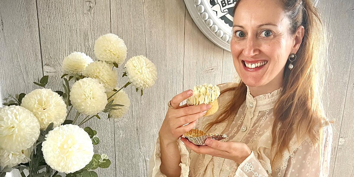 Smiley, blonde lady in a cream blouse holding a cupcake taken from a large flower-like cupcake basket.