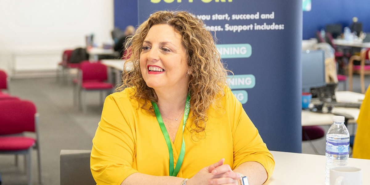 Happy, smiling woman in a yellow dress with shoulder length curly hair. In the background is an office.