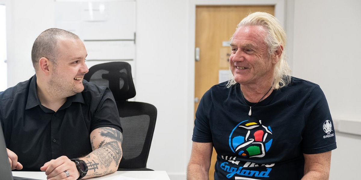 Two man sitting done chatting, looking happy. Both men are wearing black - one is wearing a black shirt with visible tattoos and the other has blonde long hair with a black football t-shirt.