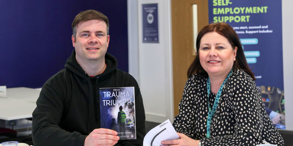 Man and woman sitting down smiling at camera in an office. Man is holding a book.