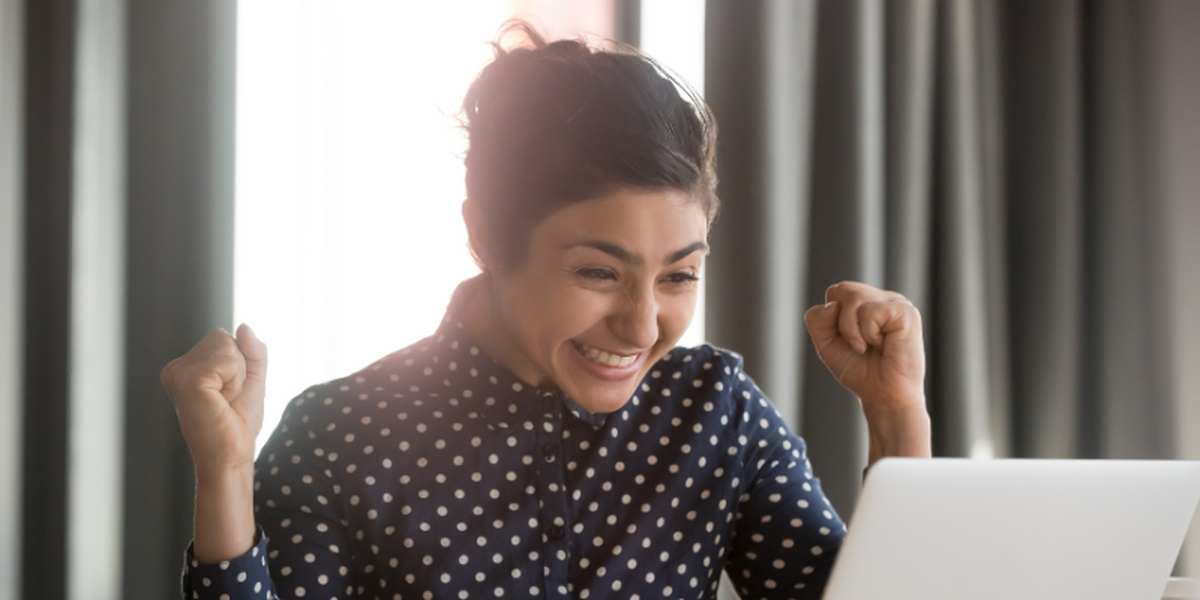 Woman in a polka dot shirt with brown hair tied back, smiling and cheering at her laptop.