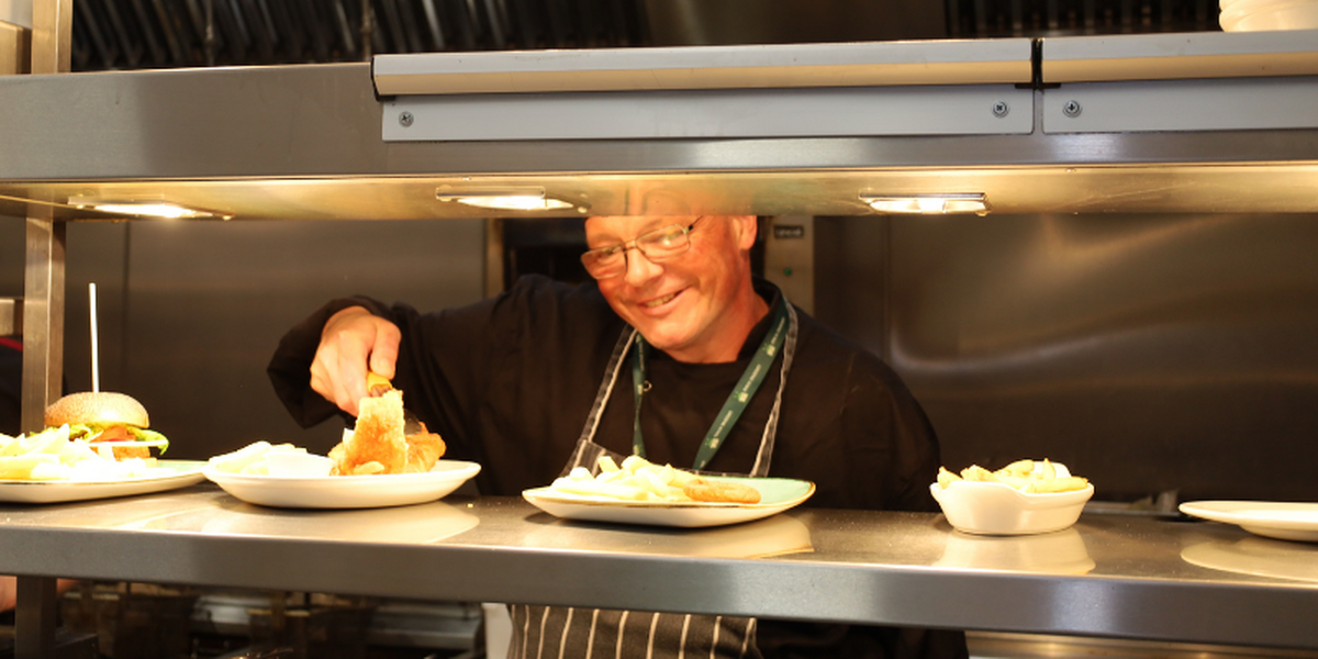 Chef on the pass, plating up food with the hot lights on. The man is smiling, wearing black and a stripey apron.