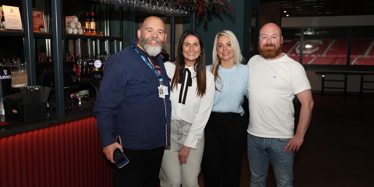 Group of two women and two man standing side by side, huddled in for a photo. All are dressed casually and smiling. Bar in the background. 