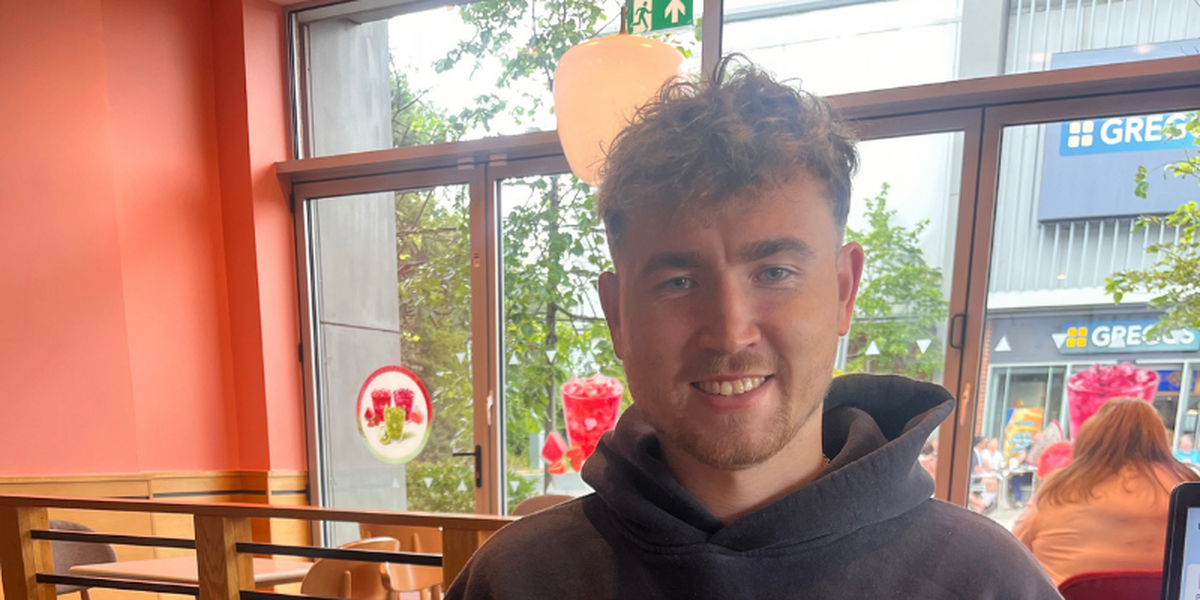 Headshot of a young man in a busy cafe decorated in orange and red. The man is wearing a hoody and has short hair.