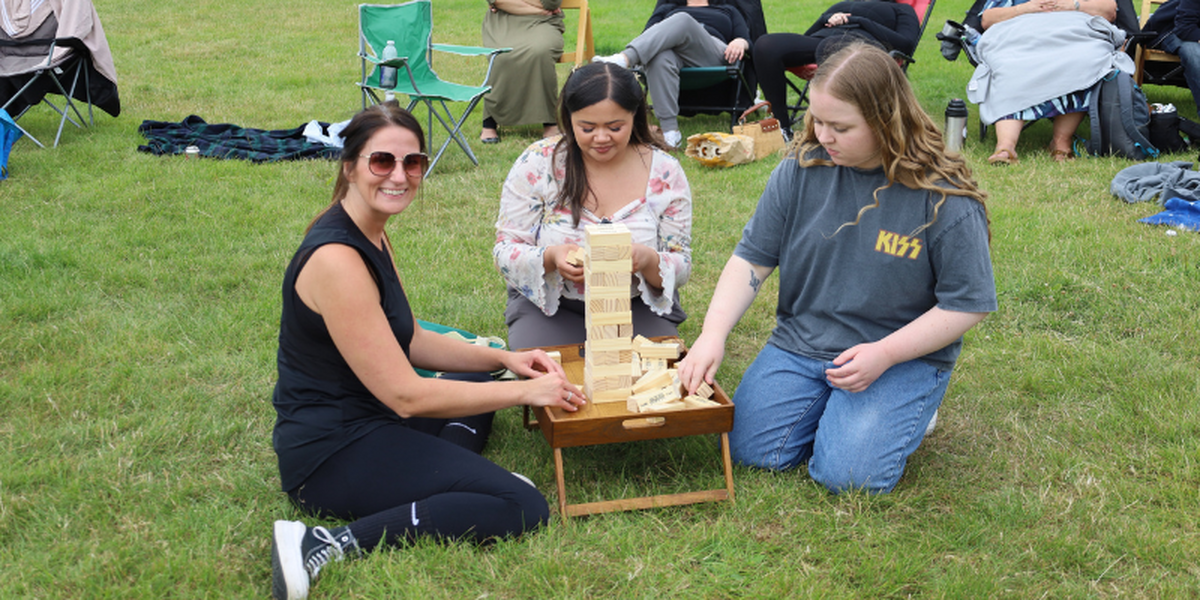 Three women on the grass playing Jenga, with a girl dressed in black sports clothes smiling at the camera while the others focus on the game.