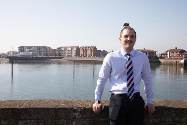 Man in shirt and tie smiling at the camera with water and buildings in background.