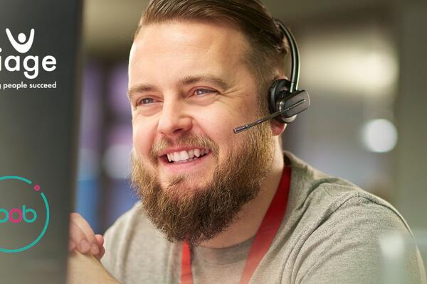 Male, bearded, customer service adviser with headset in front of computer smiling.
