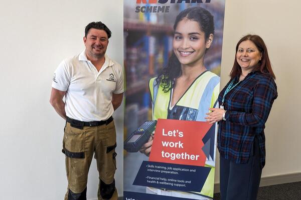 Man on the left standing beside a woman on the right with a banner promoting the Restart Scheme in the middle. Man wearing overalls and branded polo shirt promoting his handyman business. Woman in business attire.
