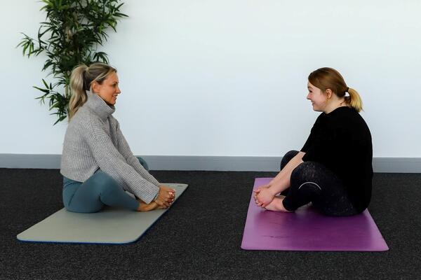 Two women sat on yoga mats with their legs crossed looking at each other.