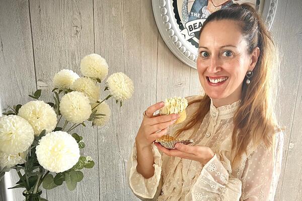 Smiley, blonde lady in a cream blouse holding a cupcake taken from a large flower-like cupcake basket.