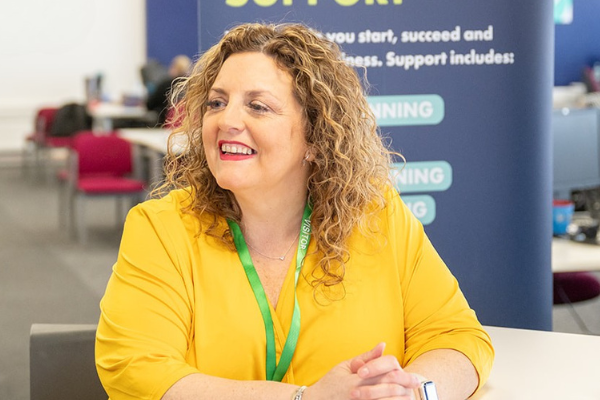 Happy, smiling woman in a yellow dress with shoulder length curly hair. In the background is an office.