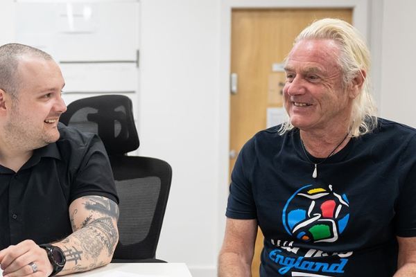 Two man sitting done chatting, looking happy. Both men are wearing black - one is wearing a black shirt with visible tattoos and the other has blonde long hair with a black football t-shirt.