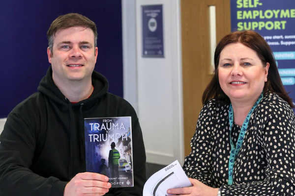 Man and woman sitting down smiling at camera in an office. Man is holding a book.