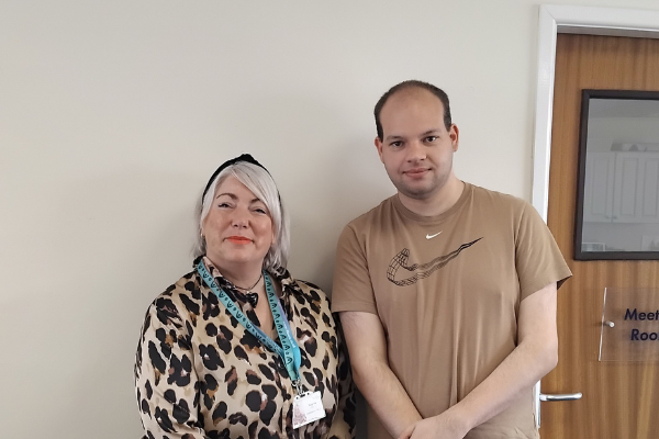 Woman with blonde hair, black hairband and leopard print shirt standing next to man with a beige Nike t-shirt smiling at camera.