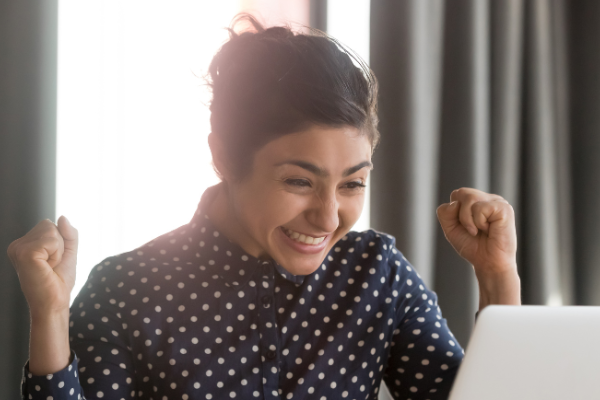 Woman in a polka dot shirt with brown hair tied back, smiling and cheering at her laptop.