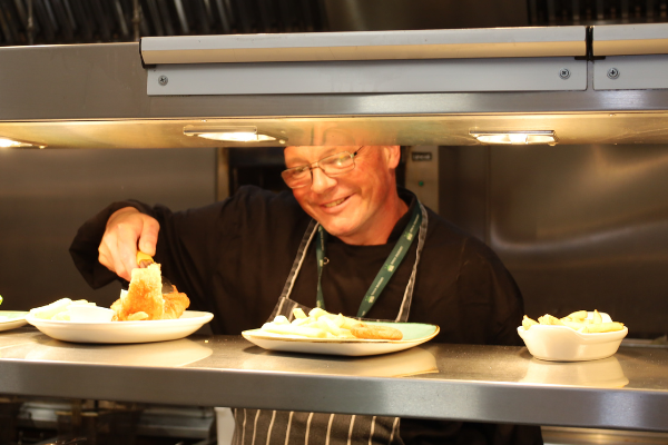 Chef on the pass, plating up food with the hot lights on. The man is smiling, wearing black and a stripey apron.