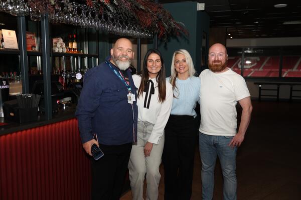 Group of two women and two man standing side by side, huddled in for a photo. All are dressed casually and smiling. Bar in the background. 