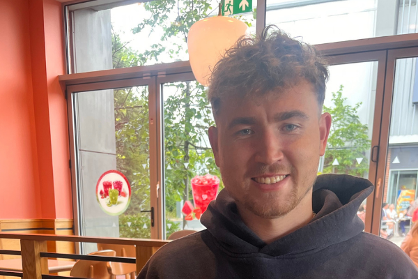 Headshot of a young man in a busy cafe decorated in orange and red. The man is wearing a hoody and has short hair.