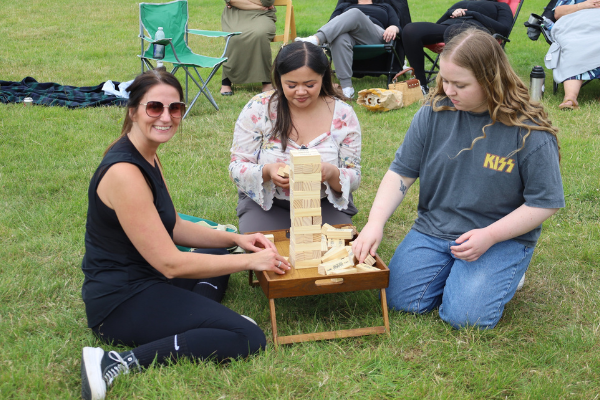 Three women on the grass playing Jenga, with a girl dressed in black sports clothes smiling at the camera while the others focus on the game.